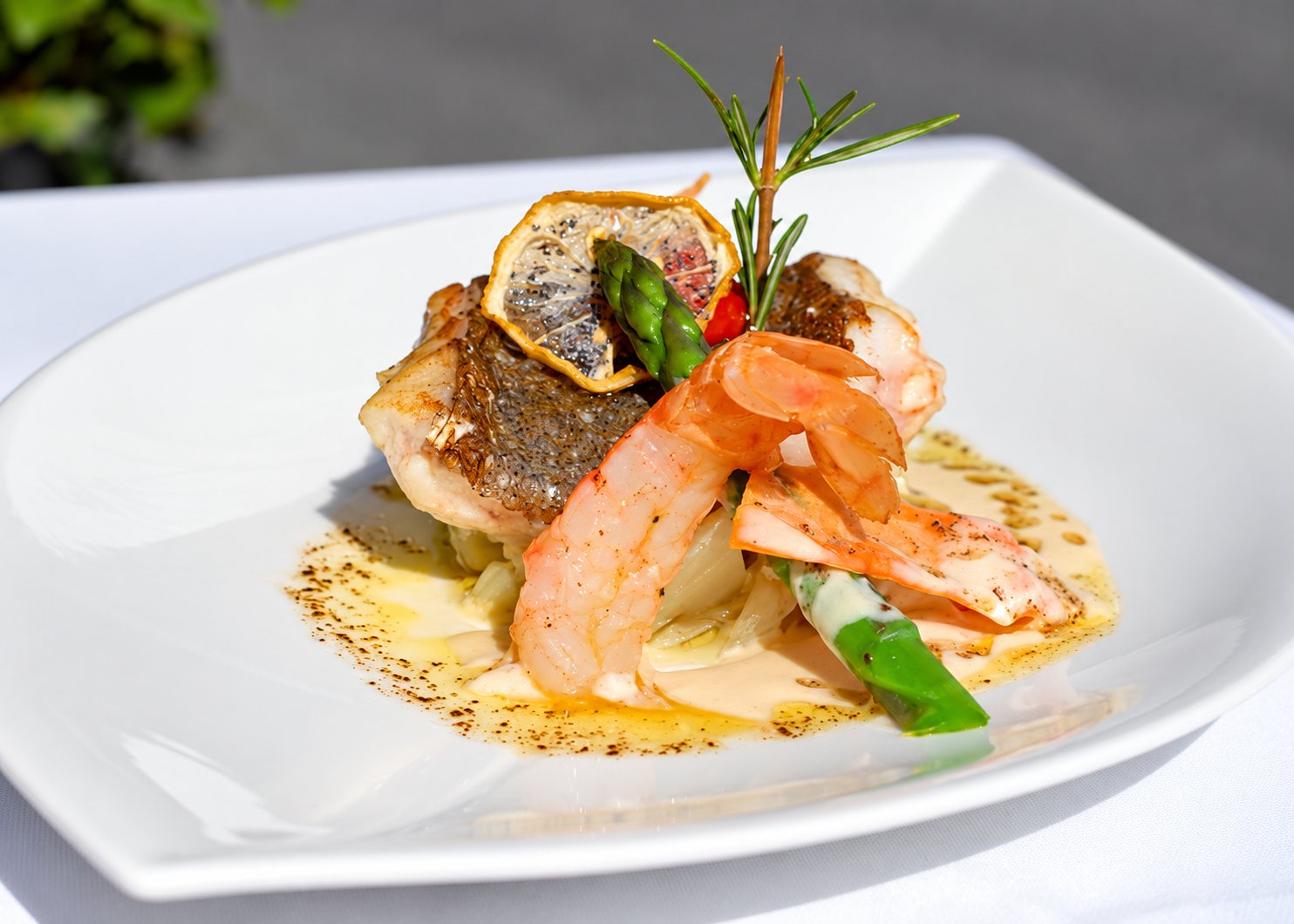 Une assiette blanche carrée présente un filet de poisson à la peau croustillante, des gambas, des asperges et une tranche de citron séché, nappés d'une sauce crémeuse et décorés d'un brin de romarin.