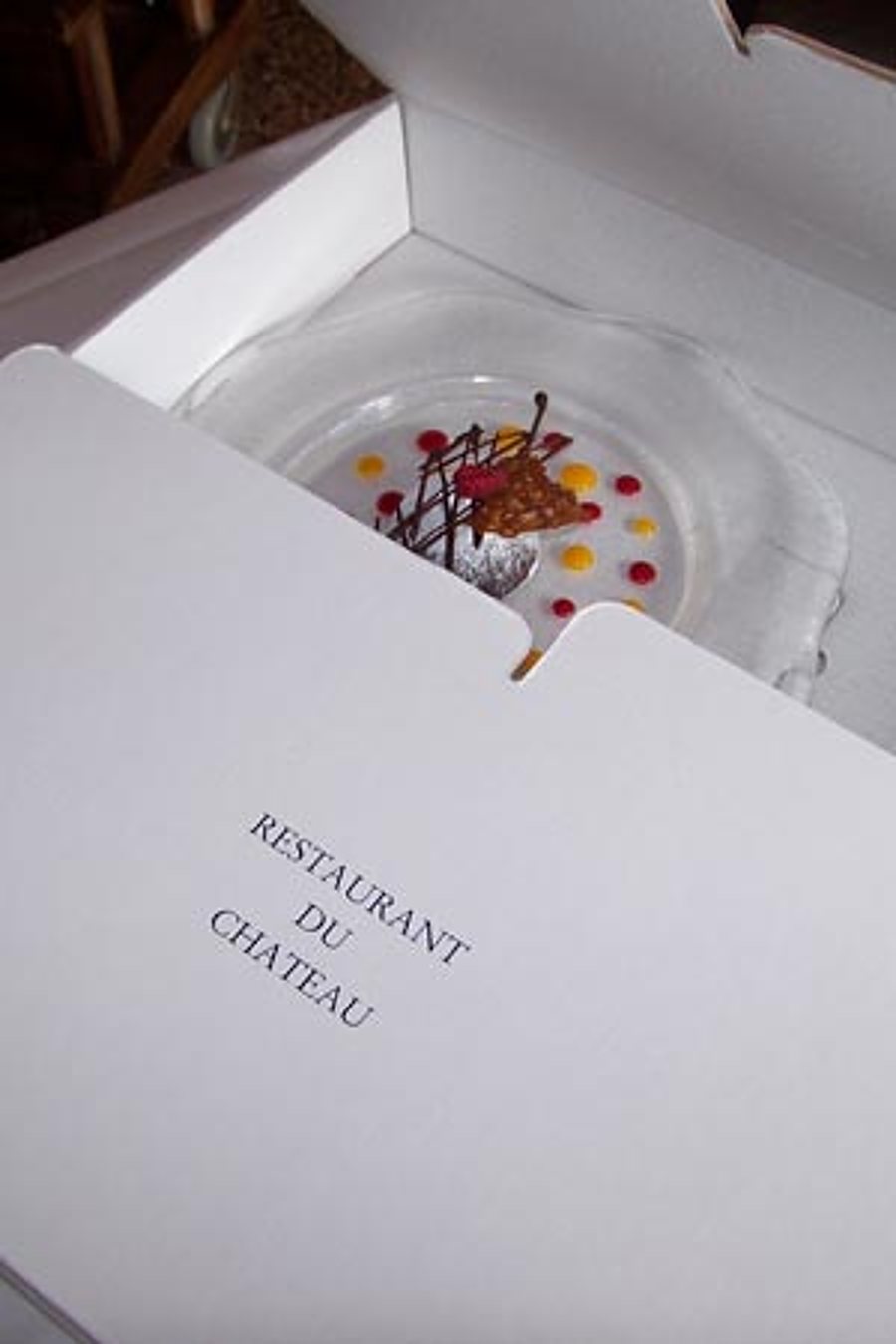 Une assiette en verre décorée d'un dessert aux points de coulis colorés et de chocolat, présentée dans une boîte blanche portant le nom Restaurant du Château.