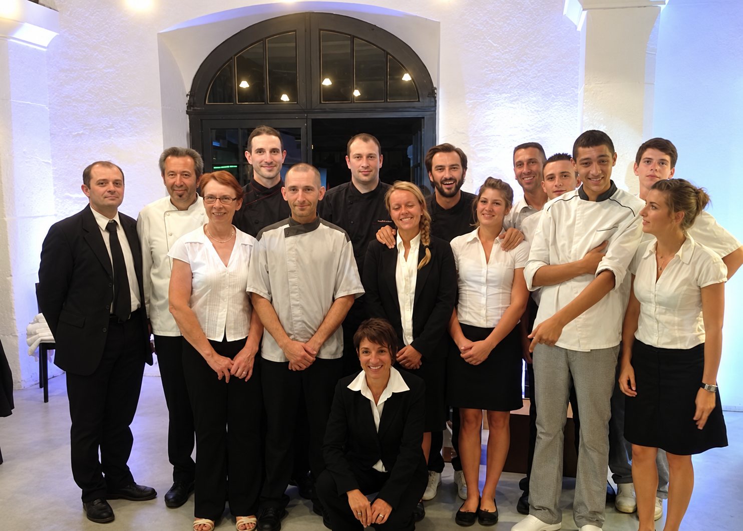 Un groupe d'une quinzaine de personnes en tenues de cuisine et de service pose devant une grande fenêtre cintrée dans une salle aux murs blancs.