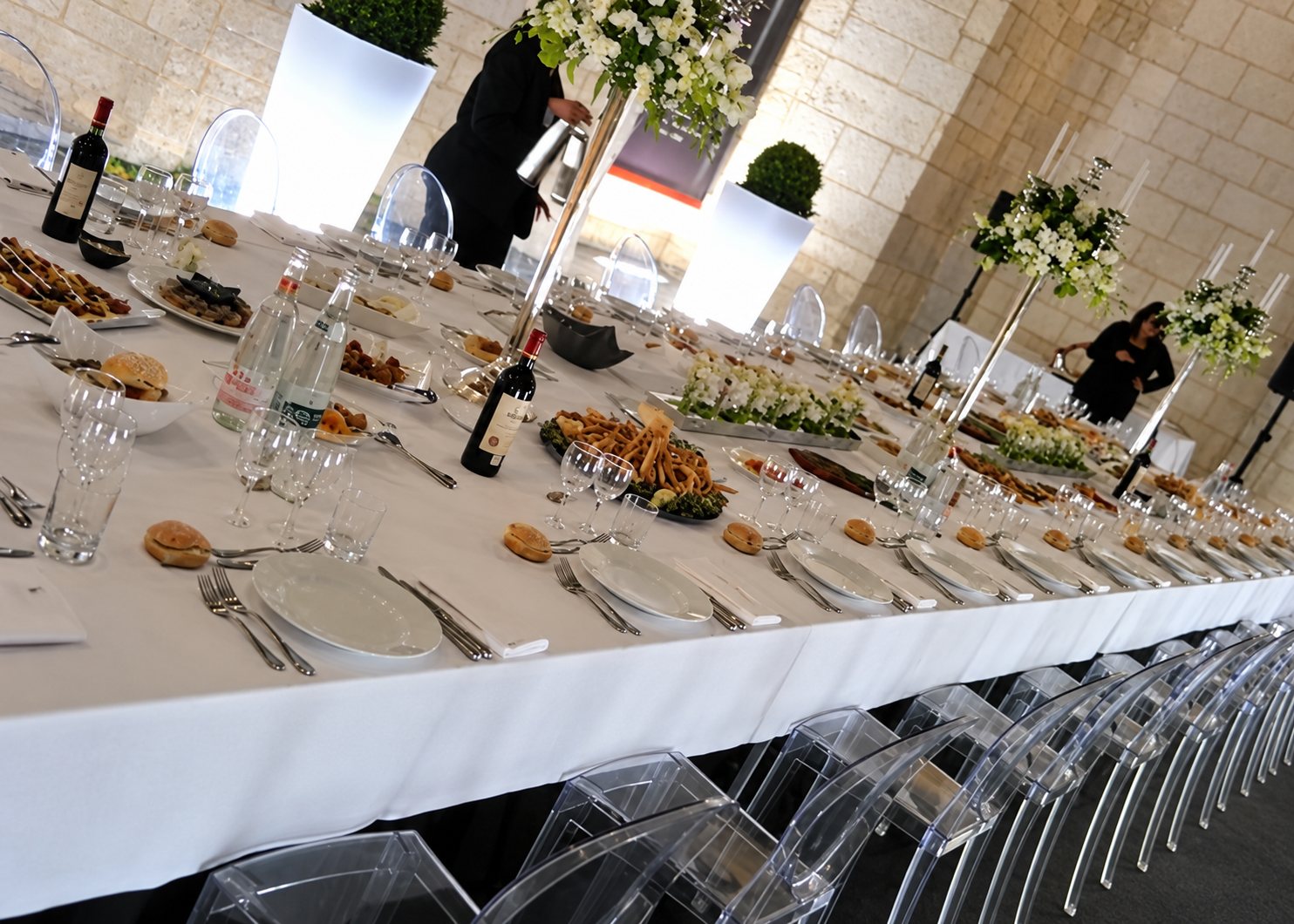 Une très longue table de réception recouverte d'une nappe blanche, garnie de plats, de verres et de bouquets, entourée de chaises transparentes dans une salle aux murs de pierre.