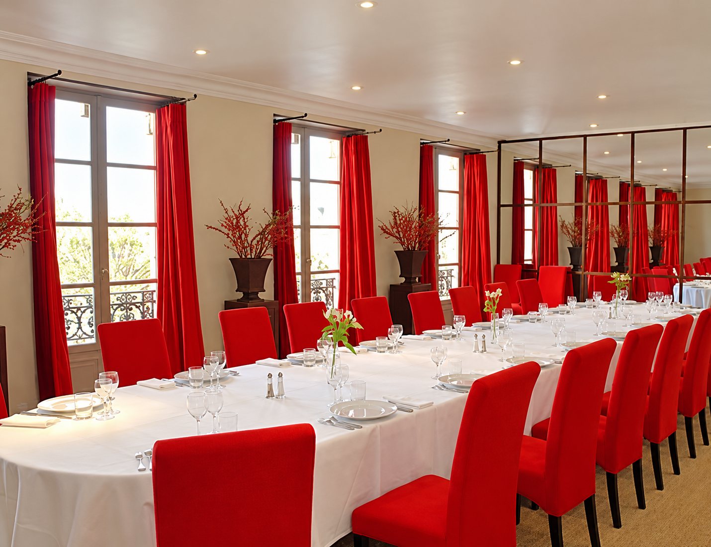 Une élégante salle de réception aux rideaux et chaises rouges, avec une longue table recouverte d'une nappe blanche et dressée pour un repas.