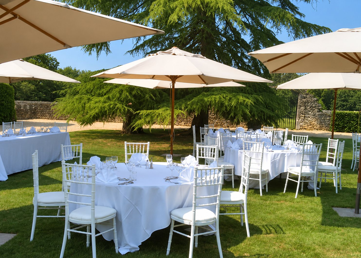 Des tables rondes aux nappes blanches, entourées de chaises blanches et abritées par de grands parasols beiges, sont dressées sur une pelouse ombragée par de grands arbres.