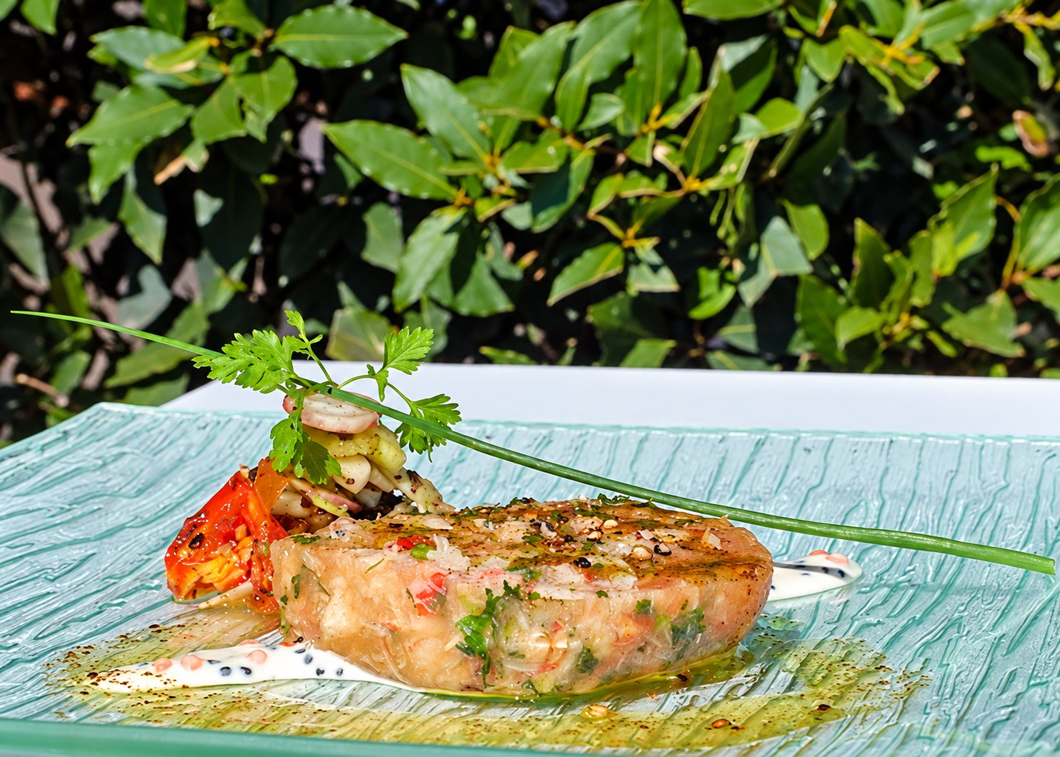 Une portion de terrine aux légumes et crustacés, garnie de cerfeuil et d'une ciboulette, est dressée sur une assiette en verre devant un feuillage vert.